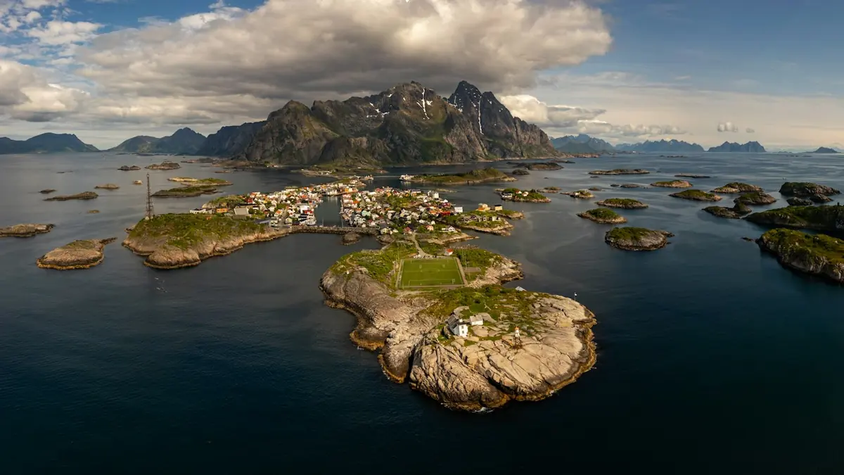 Flyfoto av et fiskevær på små øyer med fotballbane og fjell i bakgrunnen