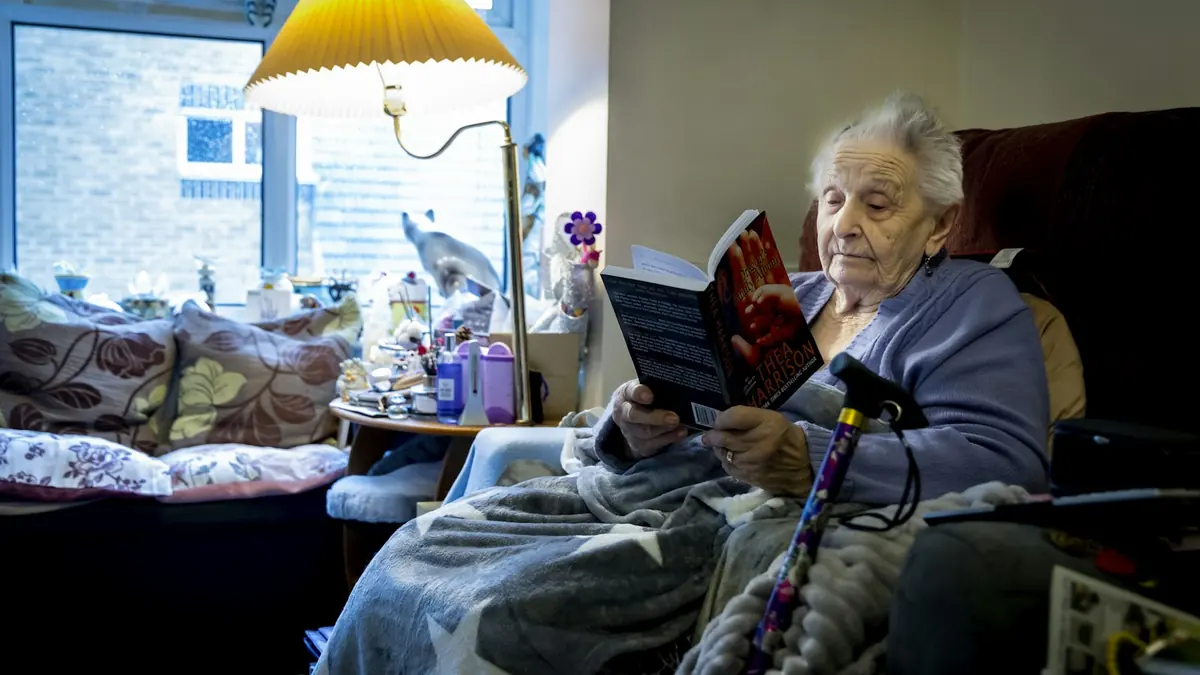 Eldre kvinne sitter i lenestolen hjemme med pledd og gangstokk og leser en bok.