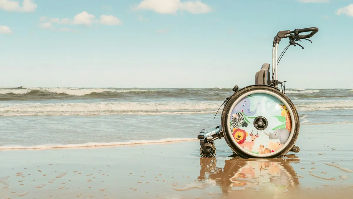 Barnerullestol med fargerike dyremotiver på hjulene står i strandkanten under blå himmel