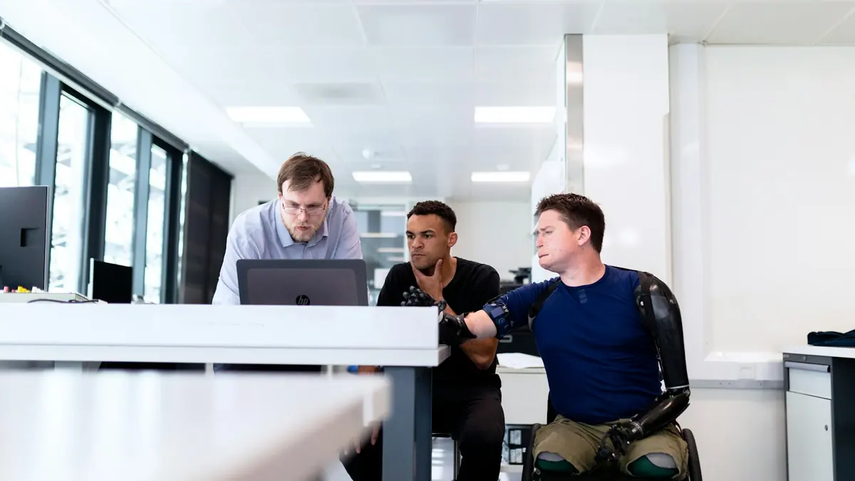 Mann med armproteser i rullestol jobber sammen med to kolleger ved en PC på et kontor.