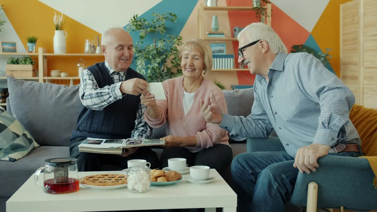 Tre eldre venner sitter i sofaen ved et bord med te og kake og ser på fotografier