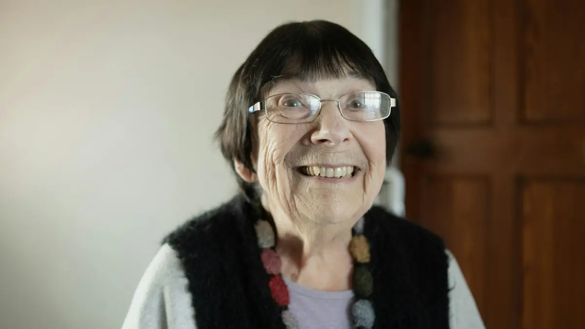 Smilende eldre kvinne med briller og strikket vest hjemme ved en tredør