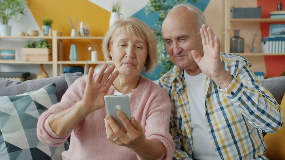 Et eldre par i sofaen vinker til noen i en videosamtale på mobiltelefonen.