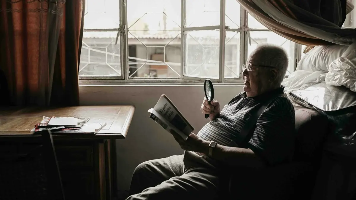 Eldre mann leser en bok med lupe i en stol ved vinduet.