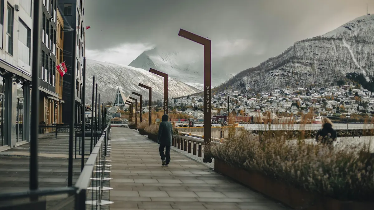 En mann går langs en kaipromenade i en nordnorsk by med snødekte fjell i bakgrunnen.