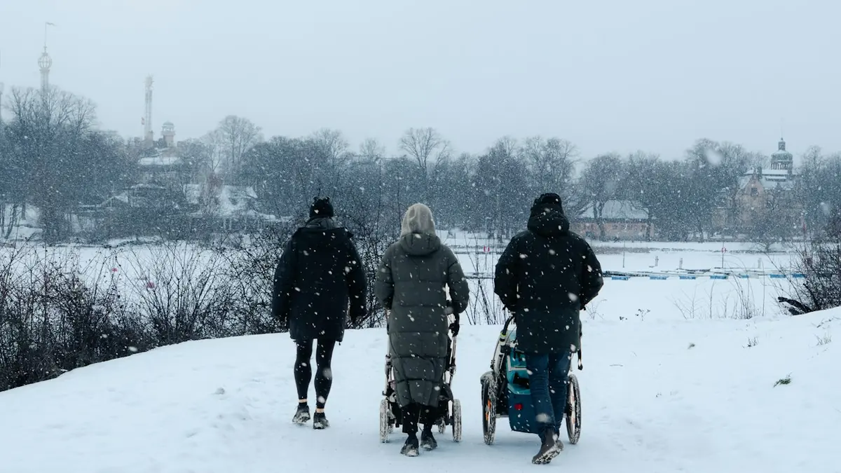 Tre voksne triller barnevogner langs en snødekt sti i snøvær, sett bakfra.