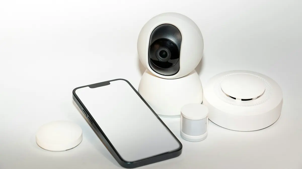 Hvite smarthusenheter – kamera, sensorer og en rund varsler – ved siden av en mobiltelefon.